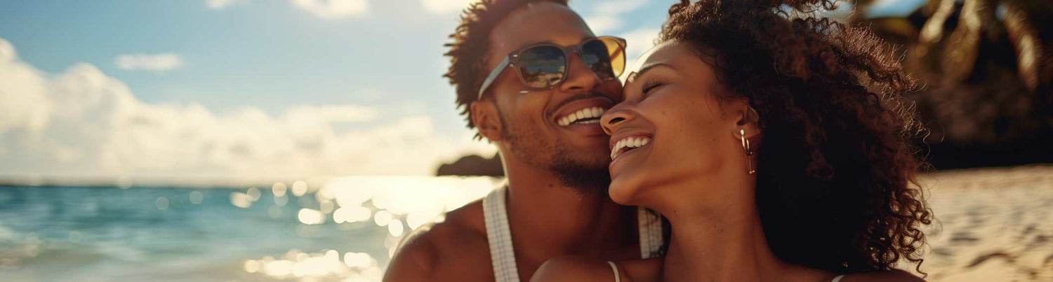 A couple sharing a loving moment on a sunlit beach, with the man smiling and embracing the woman from behind as she laughs joyfully. Their radiant health and happiness epitomize the lifestyle promoted by the AIM Companies Supplements Collection. The Good Stuff Health Shop South Africa.