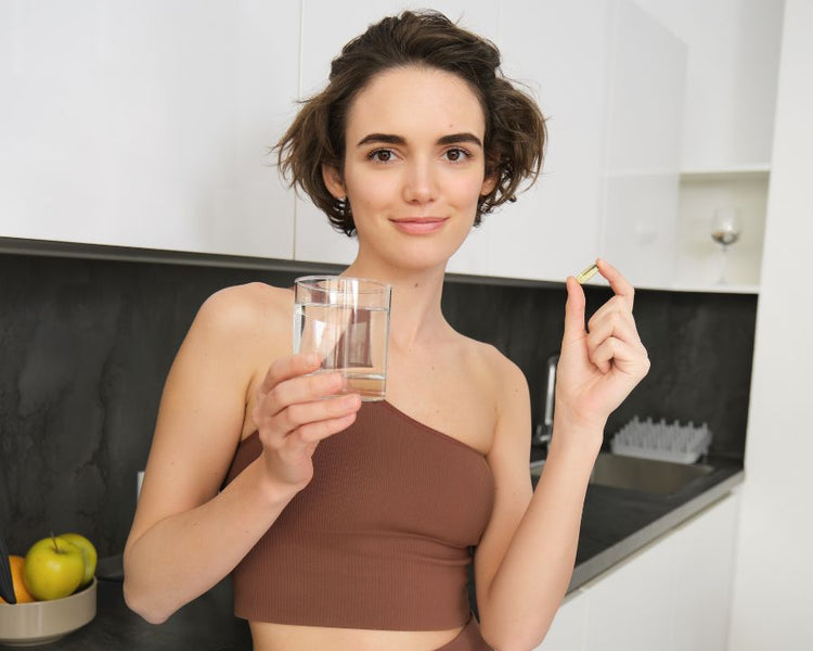 Woman in brown activewear holding a glass of water and iron supplement capsule – Best Iron Supplement For Women by The Good Stuff