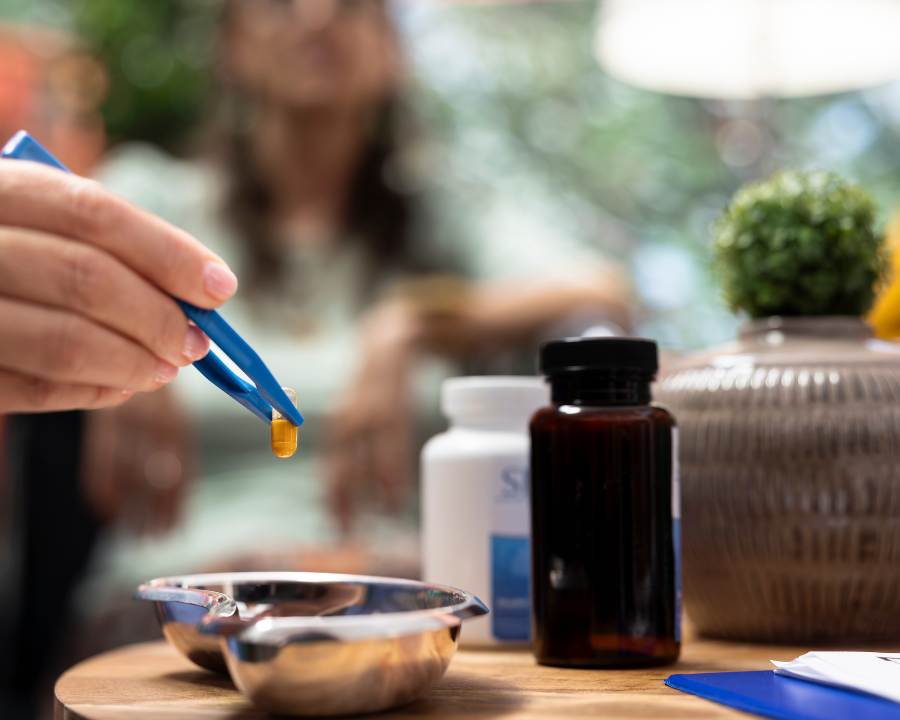 Essential Amino Acids Supplement capsule held with tweezers over a tray, with bottles and wellness items on a wooden table.