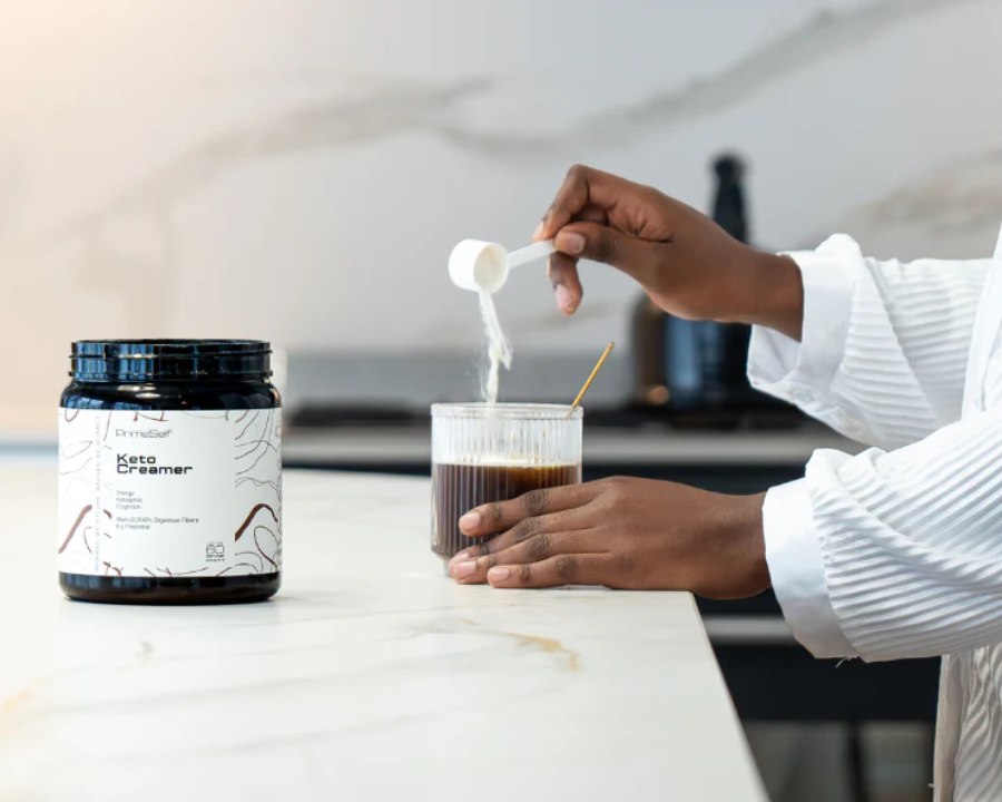 Person in a white robe pouring PRIMESELF Keto Creamer into a glass of coffee on a modern kitchen counter, promoting energy and focus.