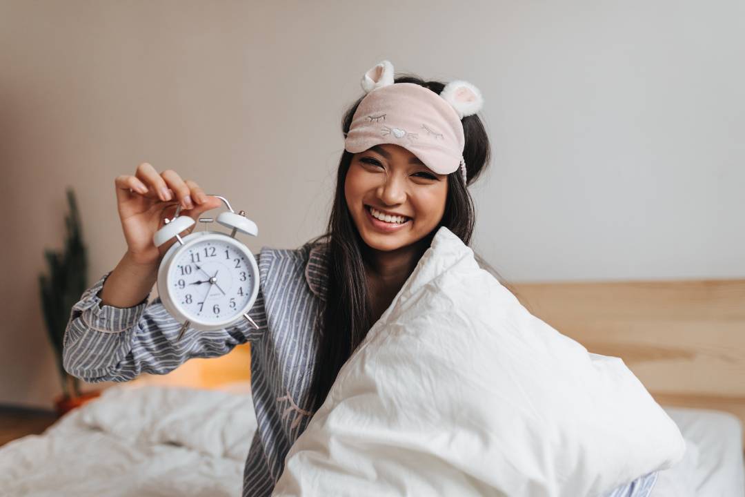 Smiling woman in pyjamas with a cute sleep mask holds a pillow and an alarm clock, highlighting sleep support supplements for better rest.