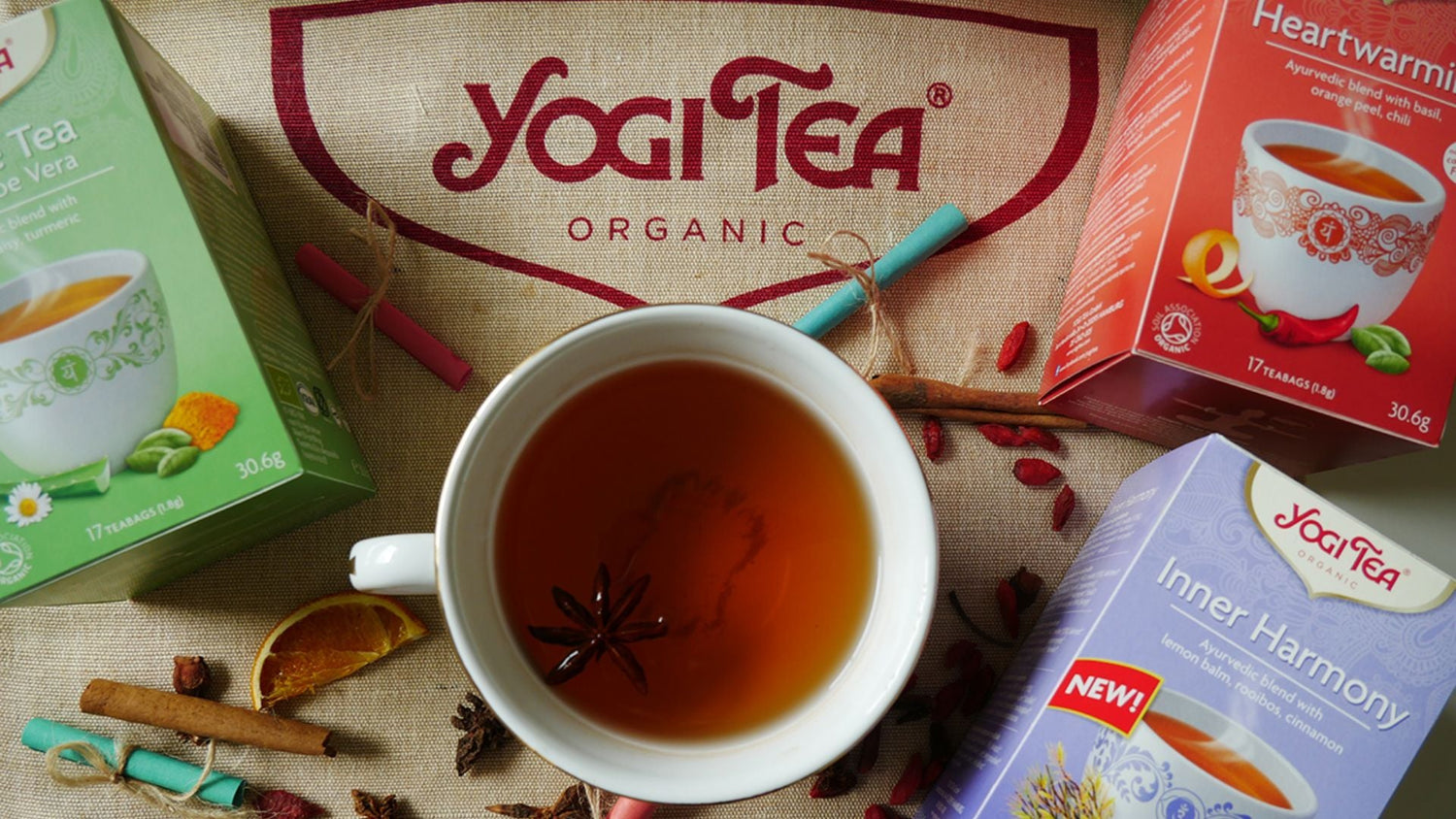 Yogi Tea organic selection with flavours including Heartwarming and Inner Harmony, surrounding a warm cup of tea and spices on a fabric mat.