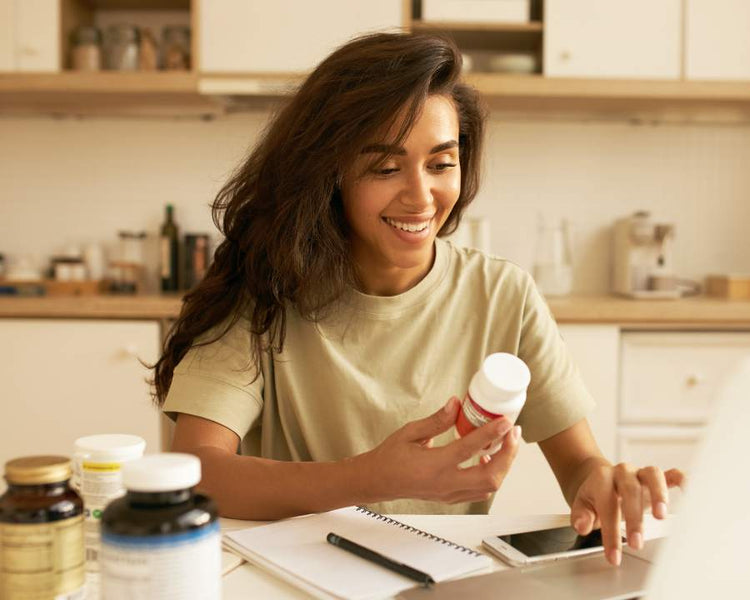 Smiling woman researching zinc supplement at kitchen table with various vitamin bottles – The Good Stuff wellness range